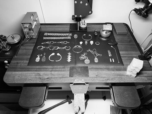 A black and white image of a jeweler's work bench, with tools and silver jewelry pieces on top. 